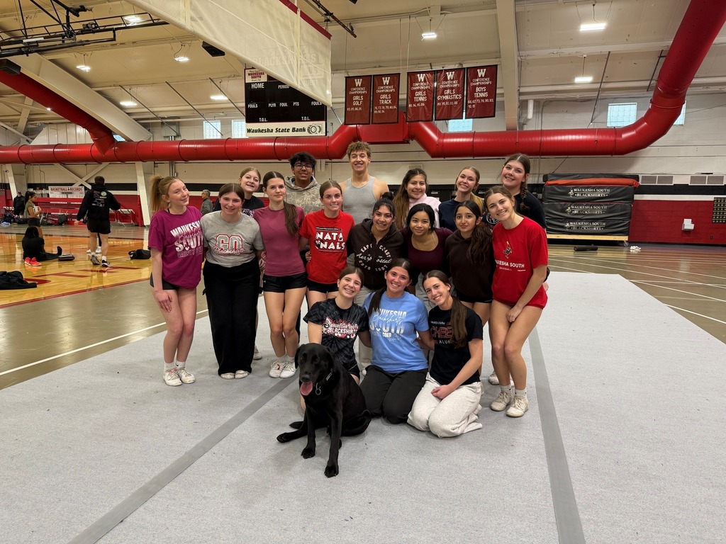Indoor court with team, coach, and dog. People in various clothing. Red pipes in background.