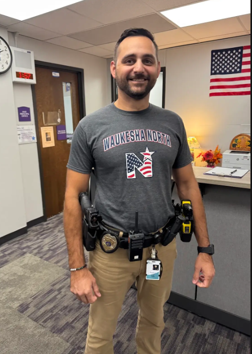 Man in a gray shirt and khaki pants with a police badge, standing in a room with a flag and a desk.