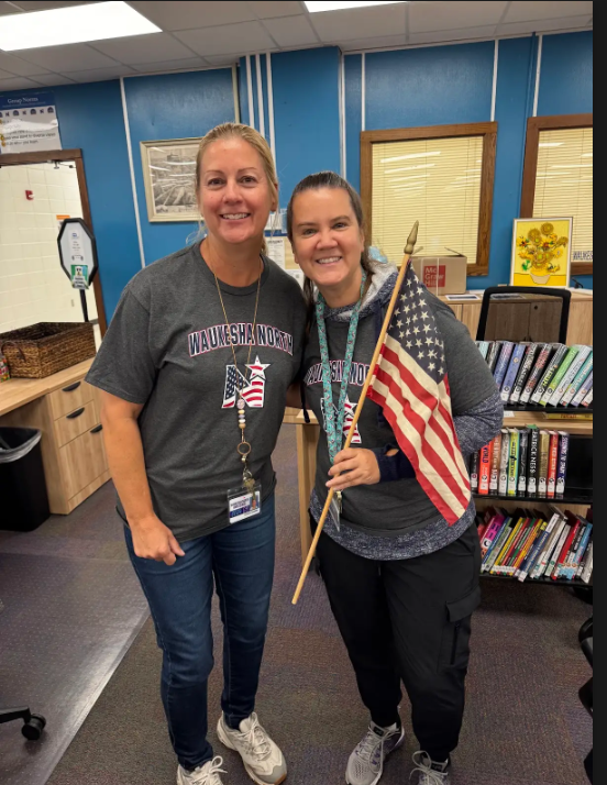 Two women in a room with bookshelves, one holding an American flag, both wearing gray shirts.