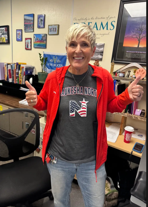 Woman in a red jacket and gray T-shirt giving two thumbs up. Behind her, a desk with a cup and other items.