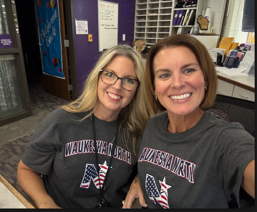 Two women with short hair smile, wearing gray shirts with "Waukesha North" text and stars. Background includes an office with a purple wall.