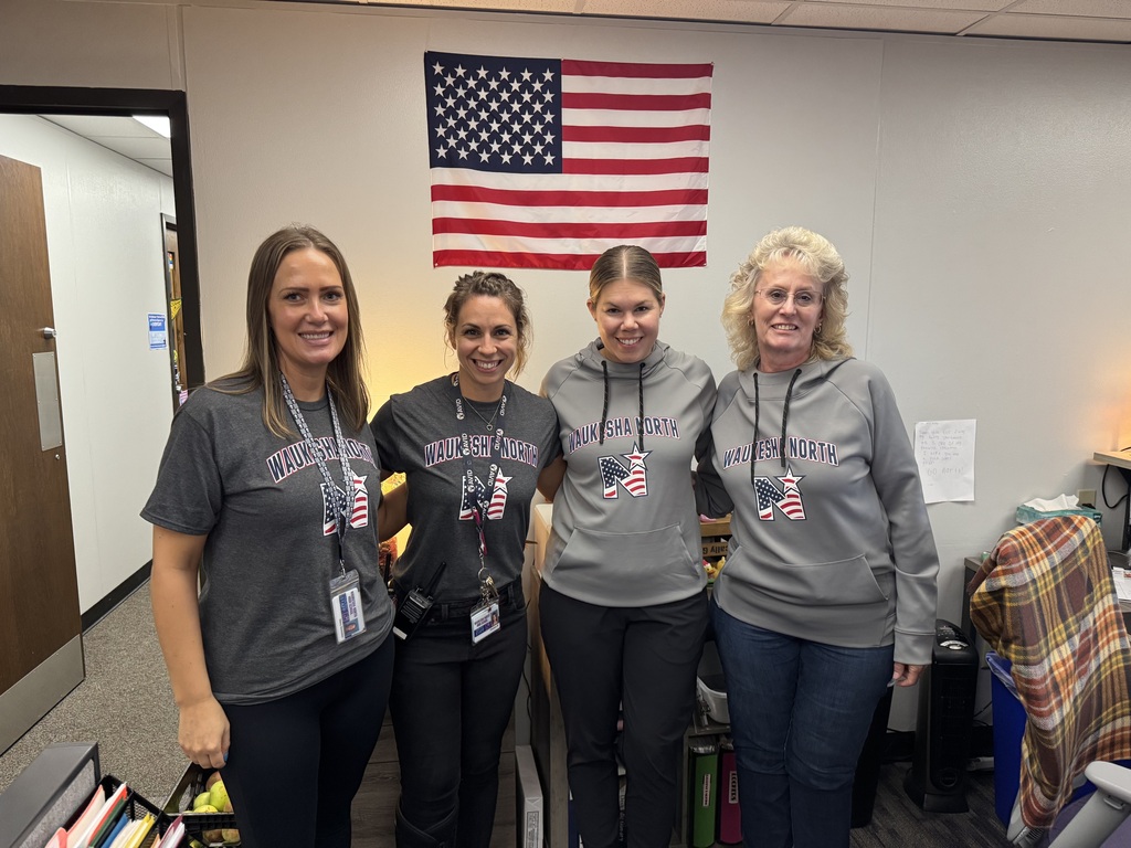 Four women stand together in an office, wearing gray shirts. Behind them is a white wall with an American flag.