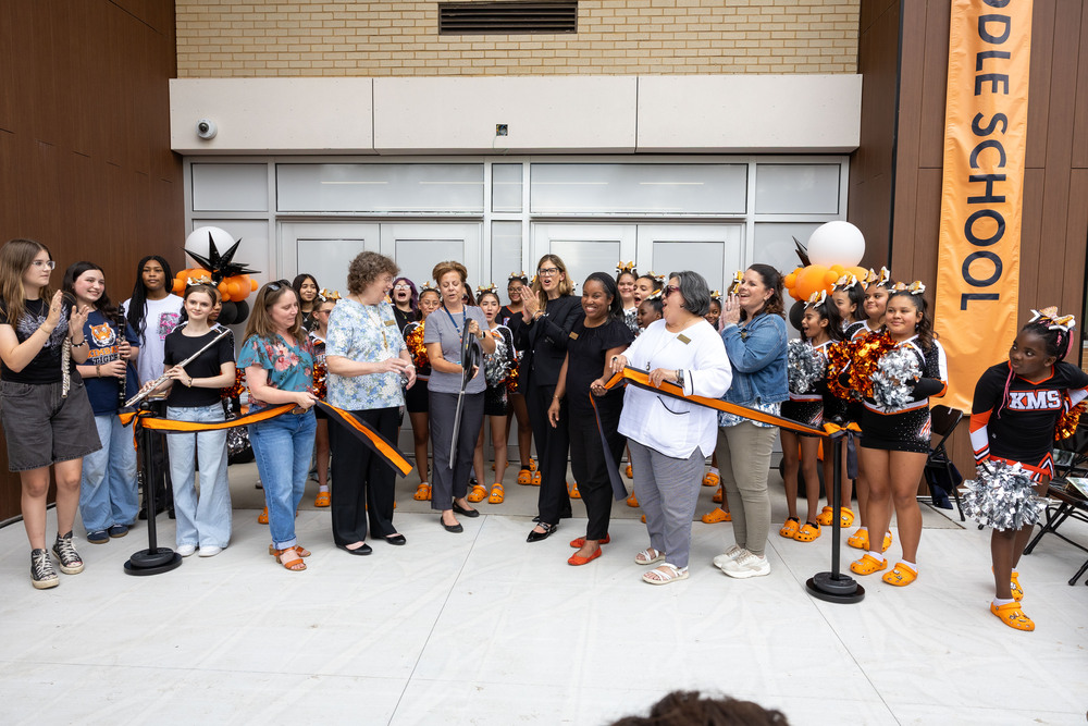 A group stands and cheers in front of a ribbon located by a school exterior
