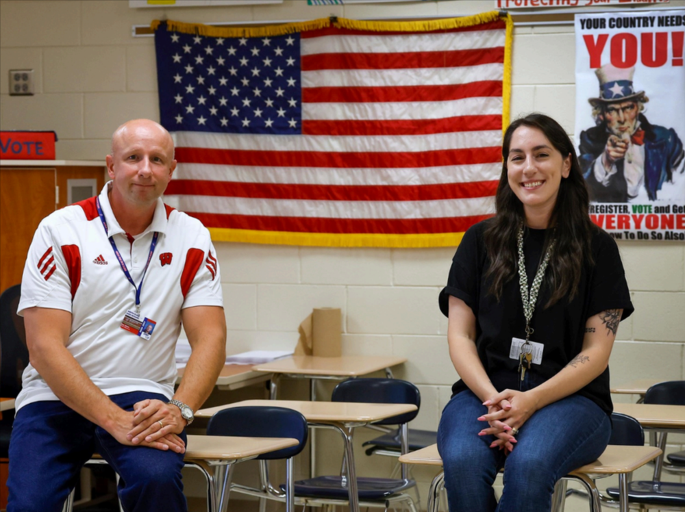 Two teachers pose in front of the American Flag in a classroom