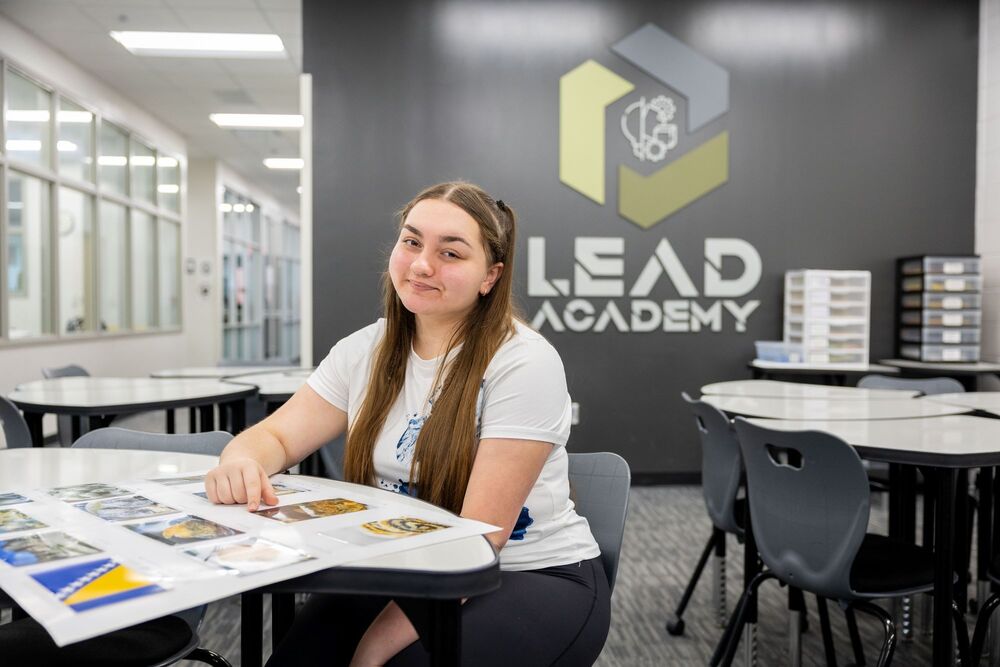 A high school student has a smile on her face sitting at a table in front of a logo that reads LEAD Academy