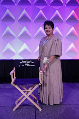 Noemi Hernandez in a gown  on a purple state by a directors chair  labeled: Kane County Educator of the Year 2026.