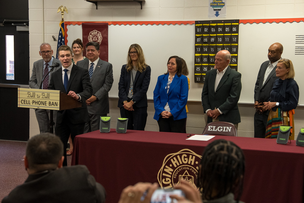 A student speaks at a podium with various adults behind him