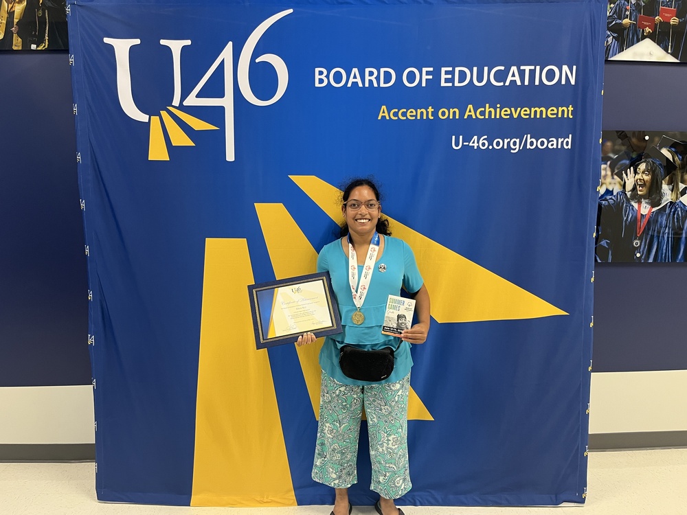 Student Rhea Raj stands in front of Accent on Achievement backdrop, holding her award