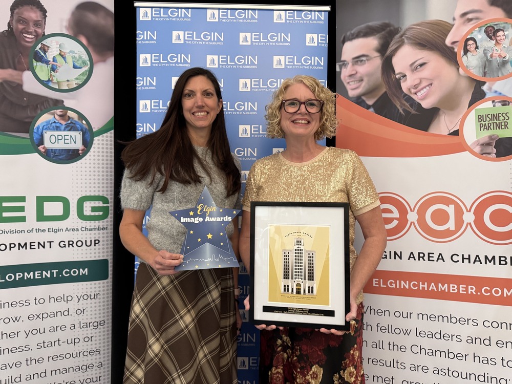 Two women stand in front of banners with smiles on their faces. One holds a cardboard star, the other an Elgin Image Award certificate.