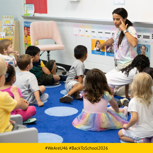 Young children sit on a rug listening to a woman in a classroom with a white board and bulletin board.