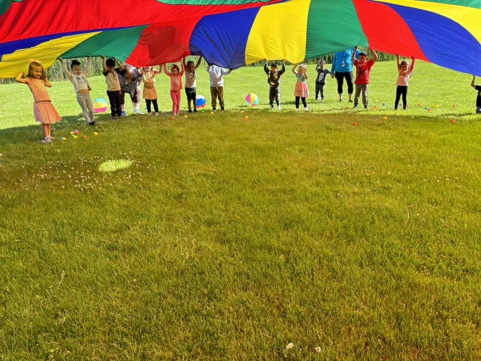 Group of children holding a large, colorful parachute outdoors on a grassy field with a clear sky.