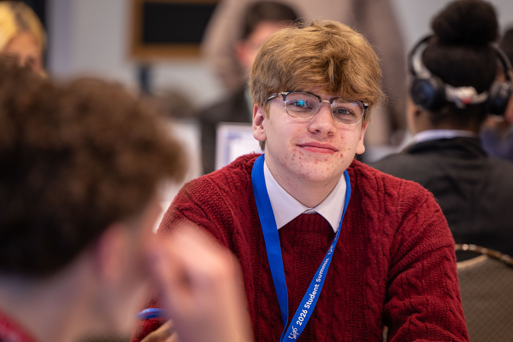 A person with glasses sits in a meeting, wearing a red sweater and a blue lanyard.