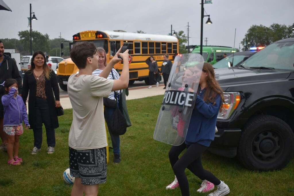 Several people stand in a park; a man points, and a woman holds a shield. A police car and school bus are in the background.