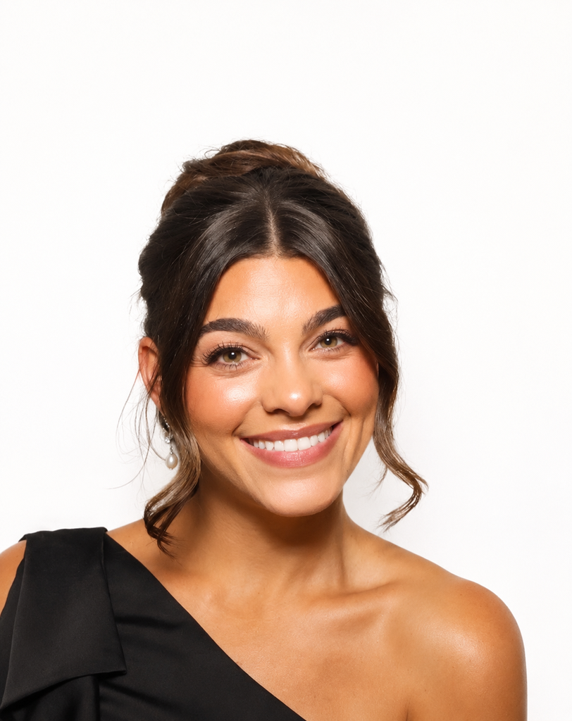 A woman with brown hair smiling, wearing a black one-shoulder dress. She has makeup and a white background.
