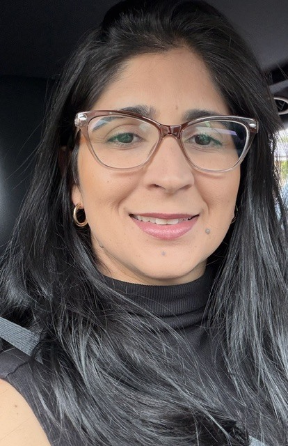 A woman with dark hair, glasses, and a black turtleneck smiles. She is in a car.