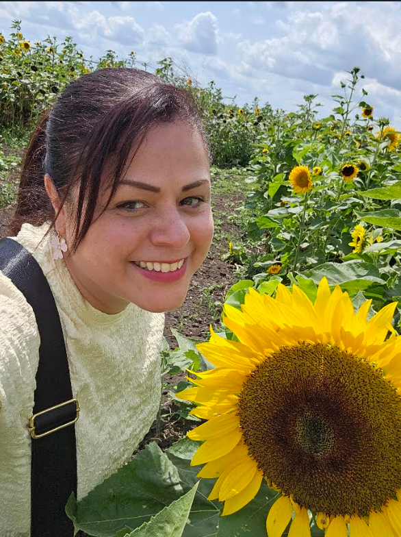 Woman wearing a white sweater holds a sunflower close to her face, smiling. Sunflowers and greenery in the background.