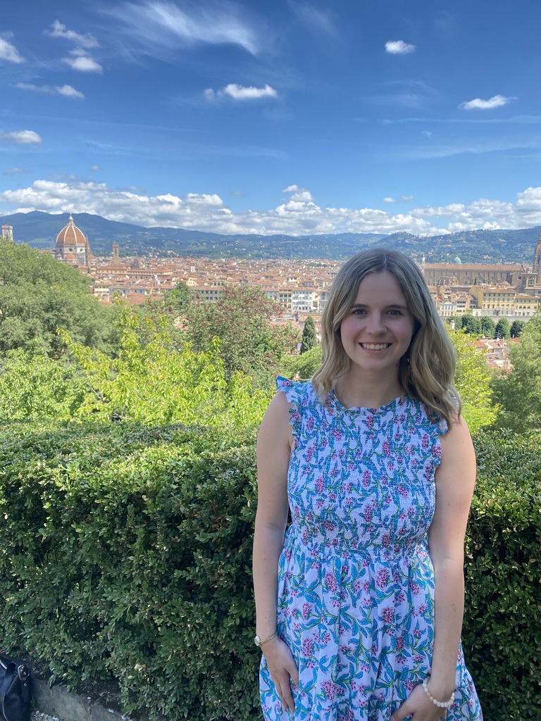 A smiling woman in a floral dress stands by a hedge, with a city skyline and hills in the background.