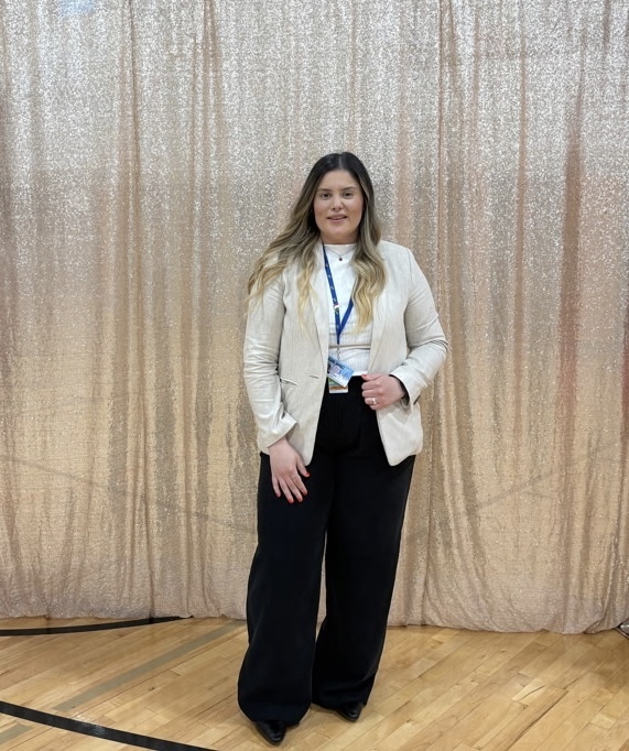 A woman in a white jacket and black pants stands in front of a golden curtain.