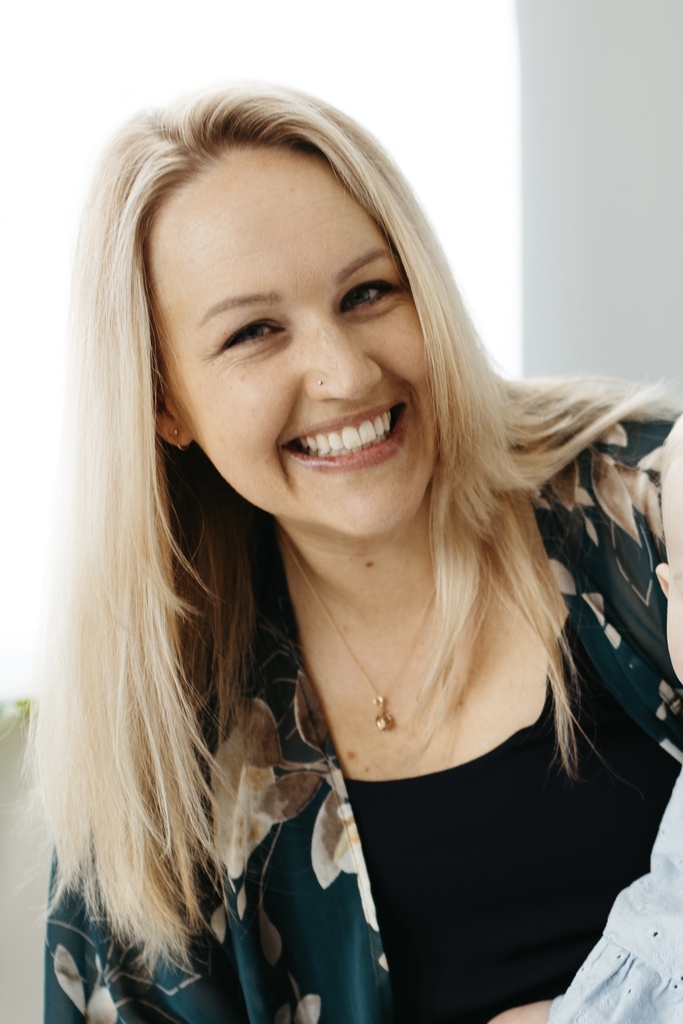 A woman with blonde hair, smiling, holds a baby. They wear a black top and a green floral jacket.