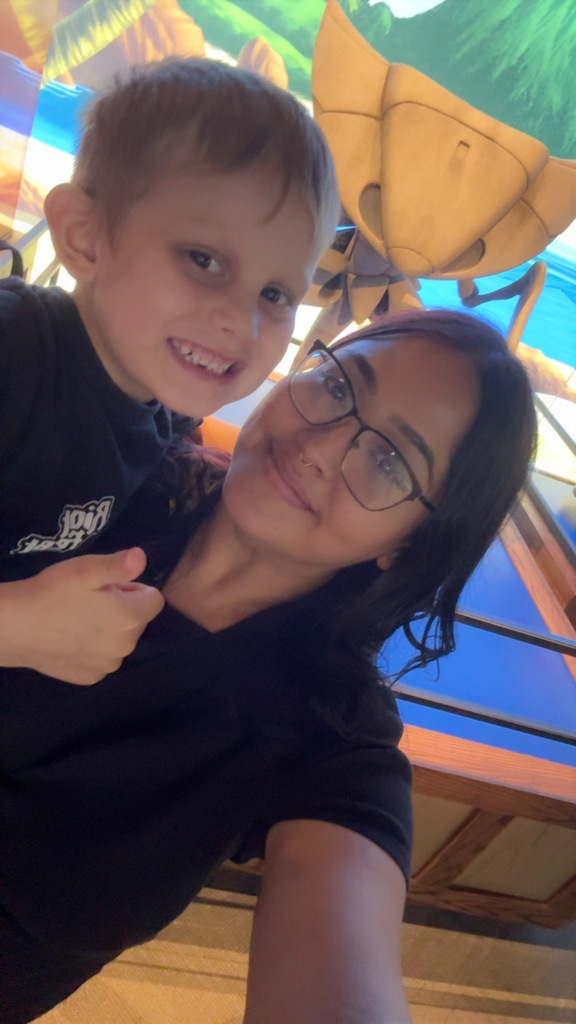 A woman and a boy are smiling in an indoor area. The boy is standing on the woman's shoulder.