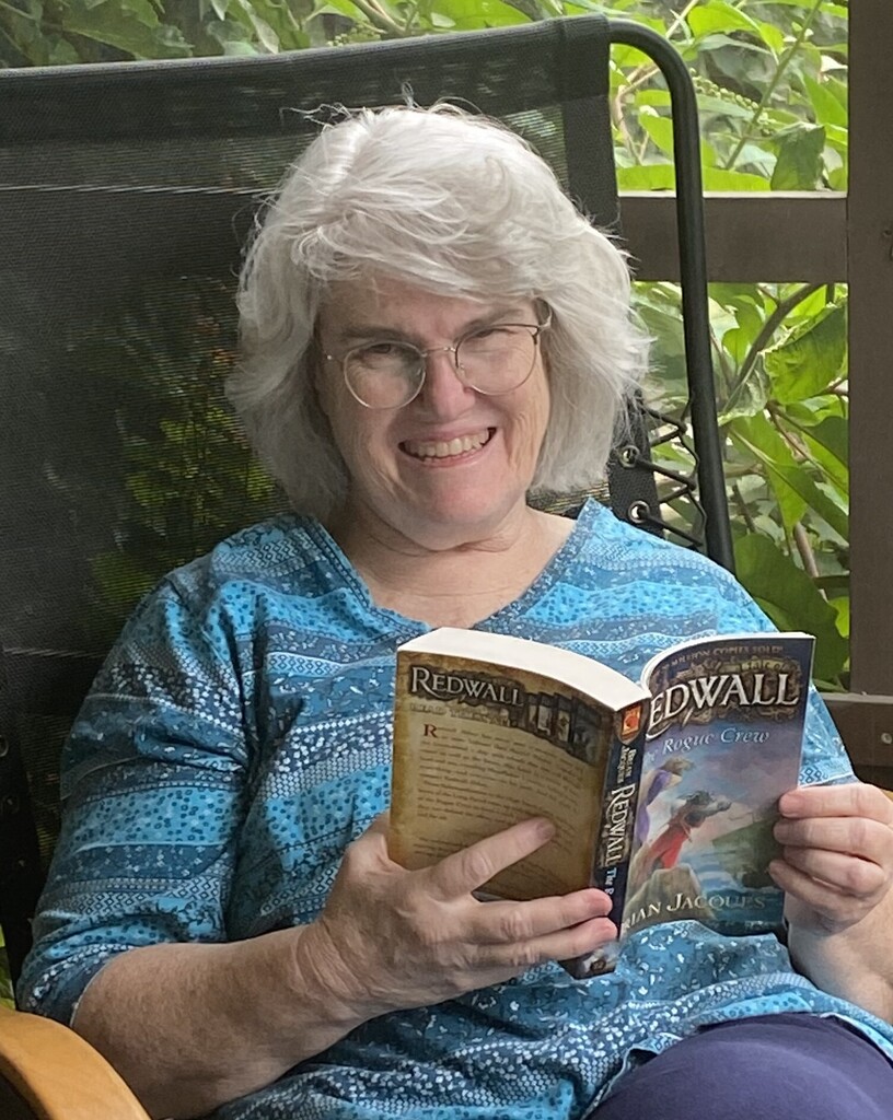 An older woman in blue sits in a chair on a porch, holding an open book. She wears glasses.