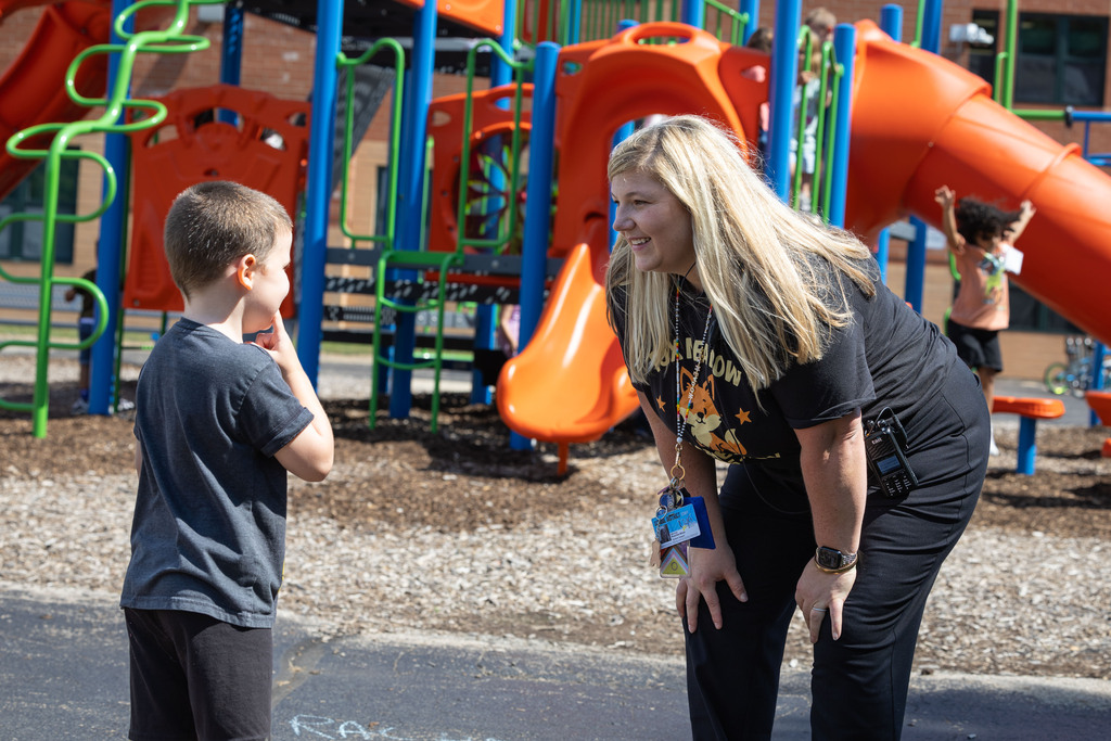 Woman with blonde hair in black shirt standing with boy in blue shirt. Background shows children playing on playground.
