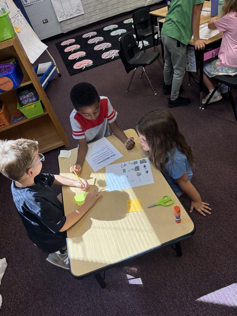 A classroom with children. Three students are seated at a table, working on an activity.