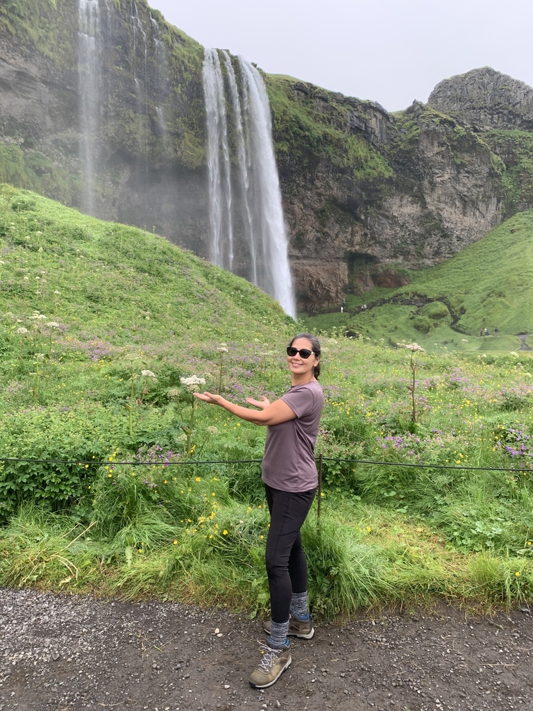 A woman in a purple shirt and dark pants stands in front of a waterfall. Behind her, a grassy hill leads to the waterfall.