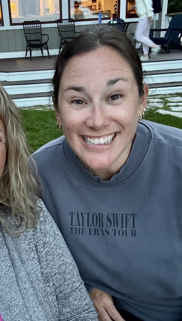 Two women smiling, one wearing a gray Taylor Swift concert shirt, on a porch with a person walking.