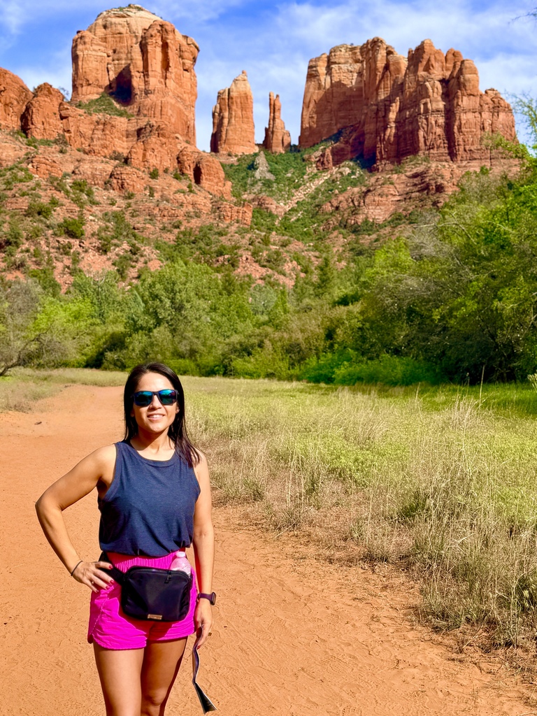 Person standing on a dirt road, wearing a blue tank top, pink shorts, and sunglasses, with a red-rock backdrop.