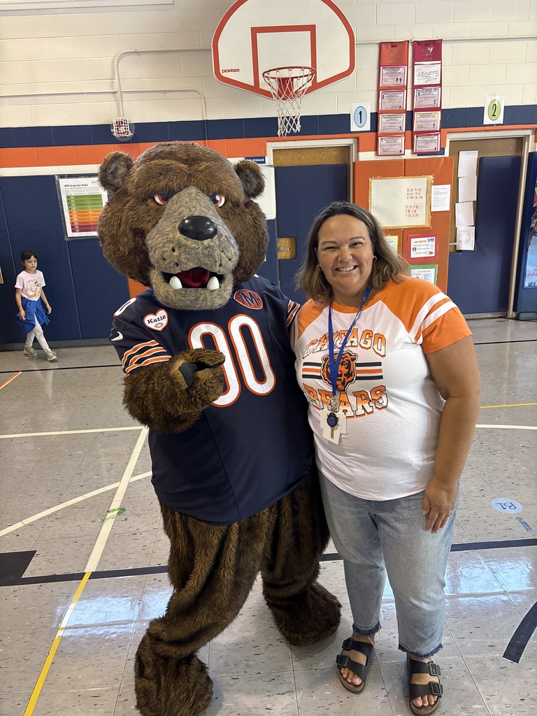 A mascot in a Chicago Bears jersey stands next to a woman in a white shirt. They stand on a basketball court.