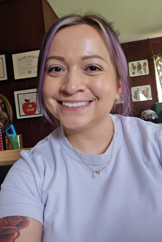 A woman with purple hair, smiling, wearing a light blue shirt, and a necklace. Background shows framed photos on a wall.