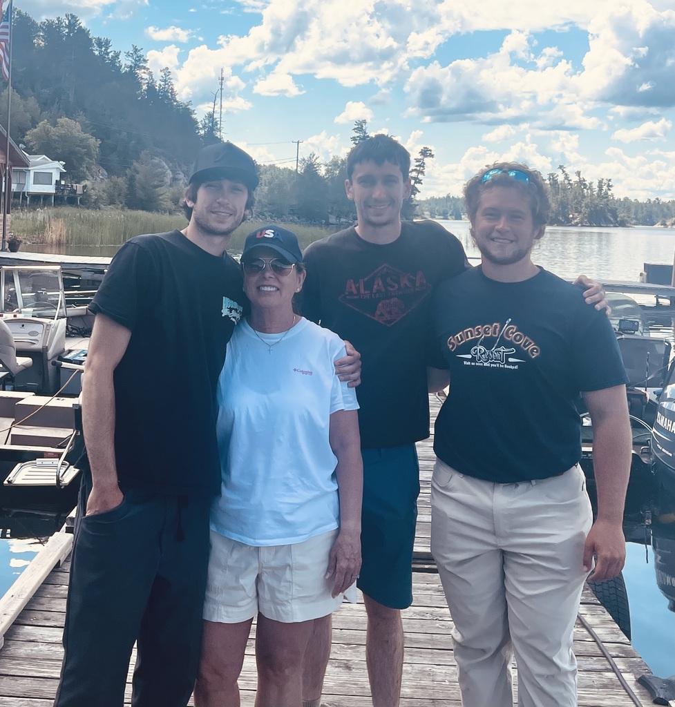 Four people on a dock by a body of water. Two men in black, one in white. Behind them, a lake with boats.
