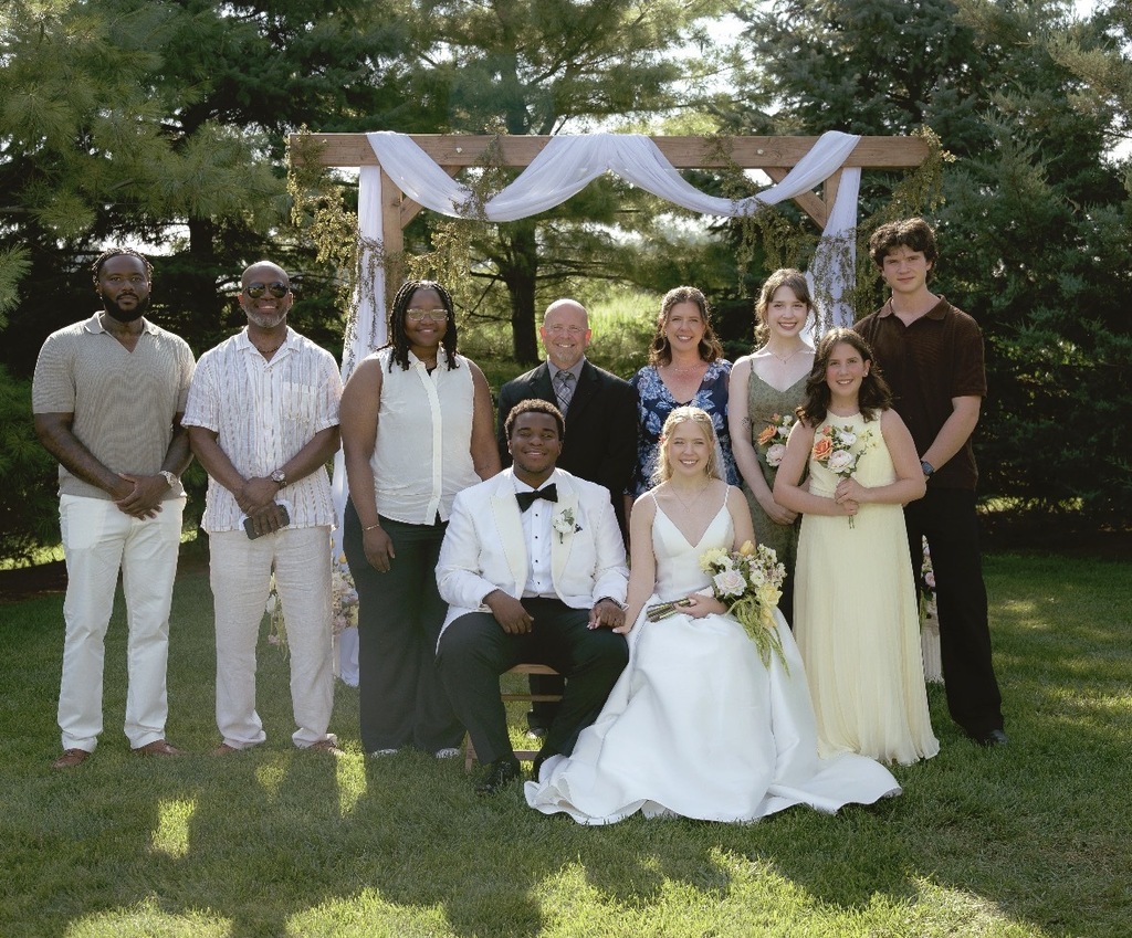 A group of people, including a bride and groom, stand in a field with a canopy arch.