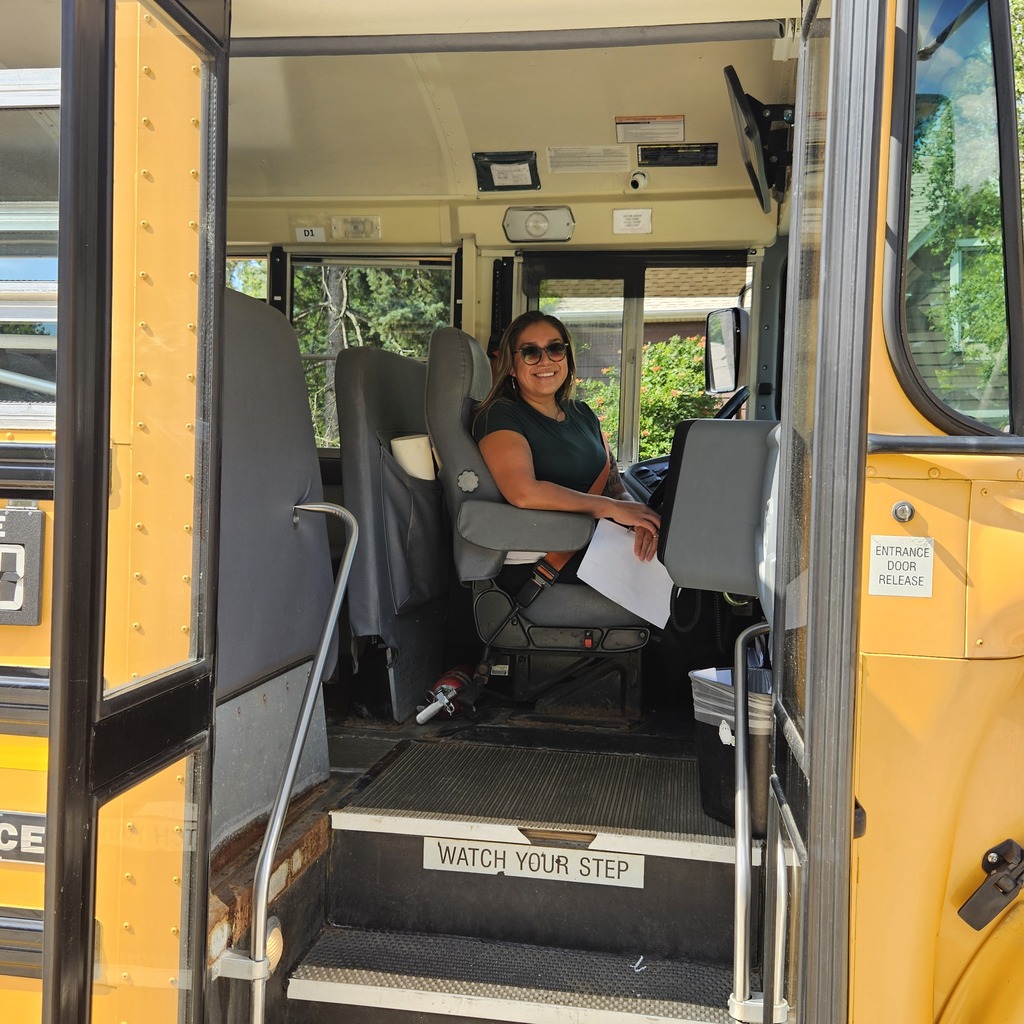 A school bus interior with a driver's seat occupied by a woman holding a paper, gray seats, and a warning sign "Watch Your Step".