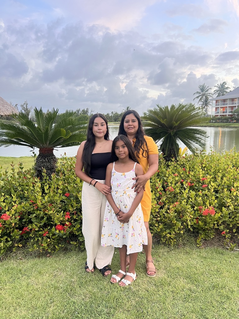 Three people in casual clothes pose in front of a lake, lush greenery, and a building.