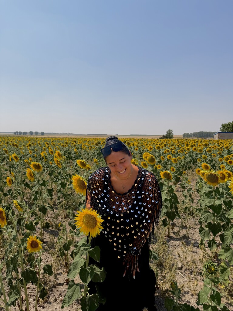 Person standing in a field of sunflowers, wearing black clothes and holding a sunflower. Clear blue sky.