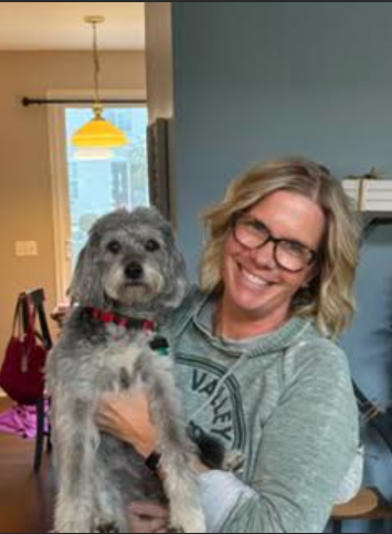 Woman with glasses holds a gray dog indoors, standing in front of a blue wall.