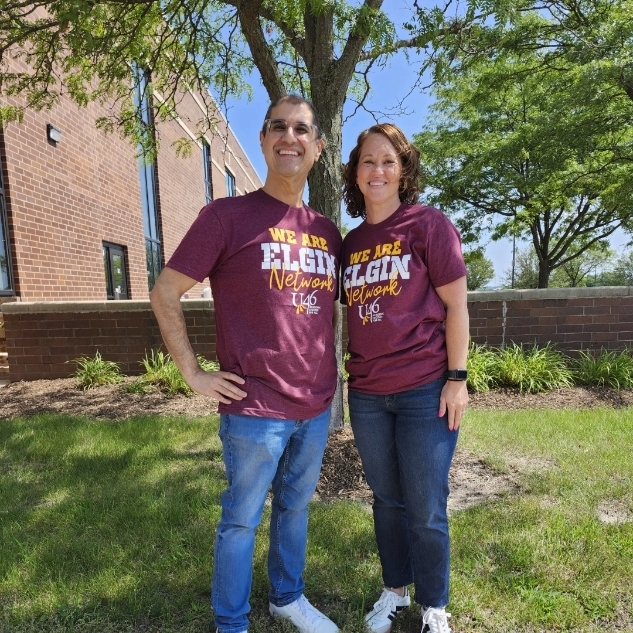 Two people stand on grass in front of a building. Both wear maroon shirts and jeans.