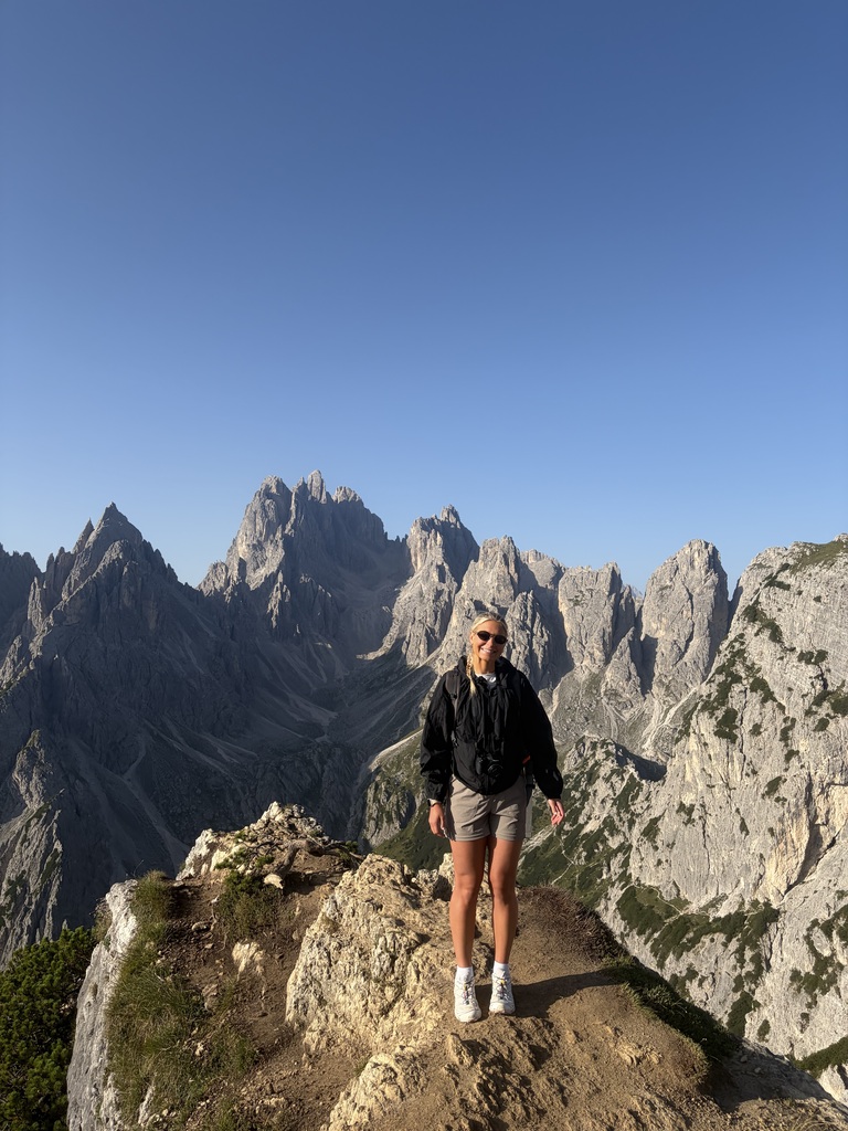 A person in a black jacket stands on a rocky cliff, overlooking a mountain range.