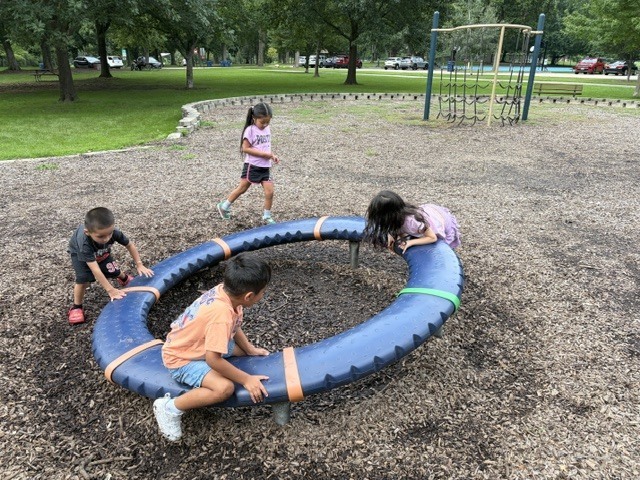 Four children play on a blue tire swing in a park, with trees and grassy areas in the background.