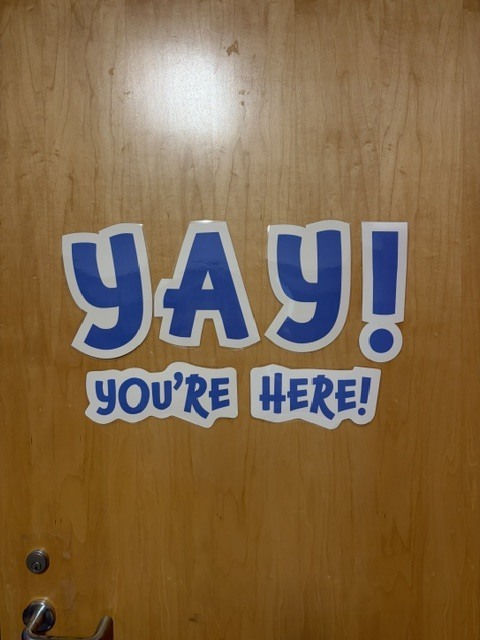 A brown door with a blue decal saying "Yay! You're Here!" in large, bold letters.