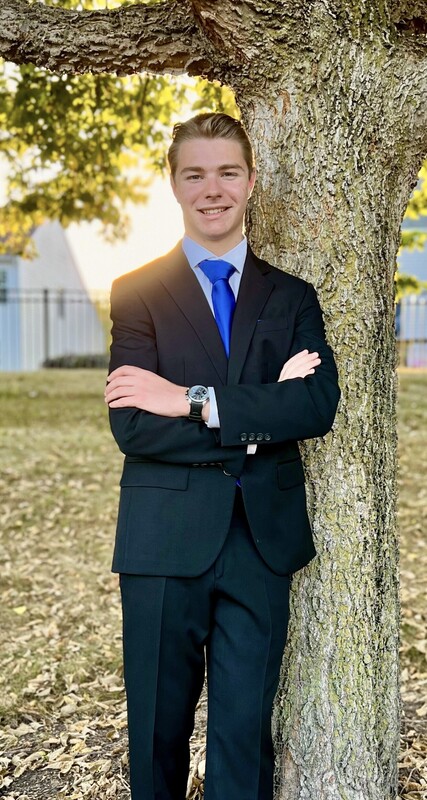 A young man leans against a tree wearing a suite