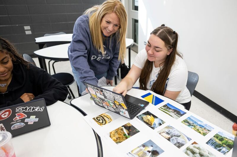 A high school student points at a laptop while a teacher with a smile on her face looks over her work