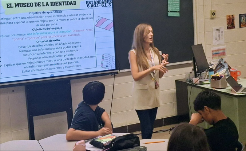 A young female teacher stands in a classroom in front of a onitor with a lesson in Spanish on it