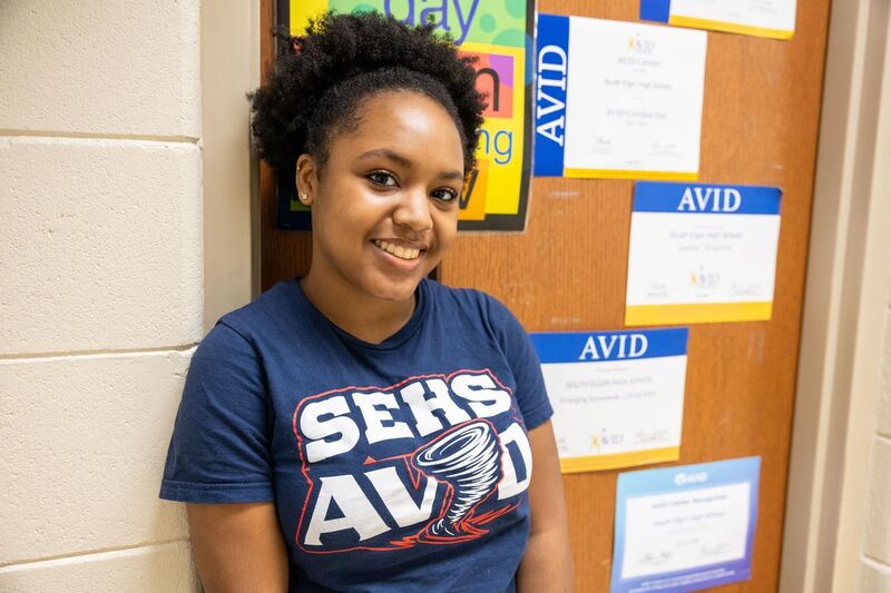 A teenage woman smiles and leans against a door. She is wearing  a blue t-shirt that reads SEHS and AVID.