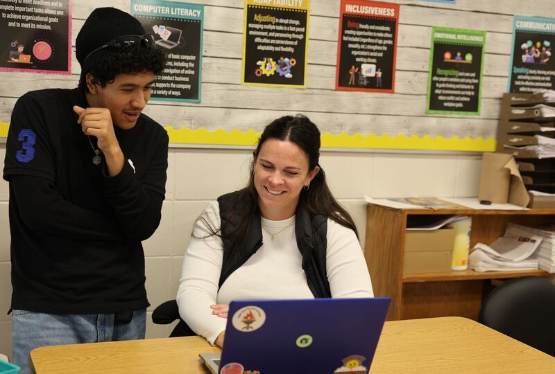 A young man smiles as he looks over his teacher's shoulder. They both are looking at a laptop.