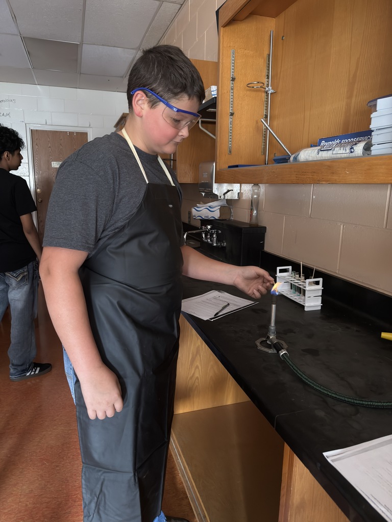 A group of students in a science classroom are gathered around lab tables wearing safety goggles. They are conducting a flame test experiment, carefully holding metal salts over a small flame. Bright colors—such as red, green, and yellow—flicker in the flames, showing the different chemical compositions. The students look focused and excited, actively participating in the hands-on activity under the guidance of Mrs. Ramos.