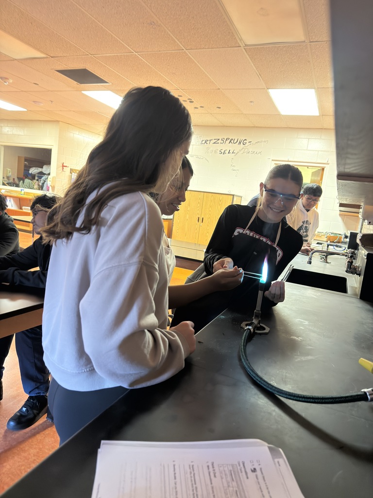 A group of students in a science classroom are gathered around lab tables wearing safety goggles. They are conducting a flame test experiment, carefully holding metal salts over a small flame. Bright colors—such as red, green, and yellow—flicker in the flames, showing the different chemical compositions. The students look focused and excited, actively participating in the hands-on activity under the guidance of Mrs. Ramos.