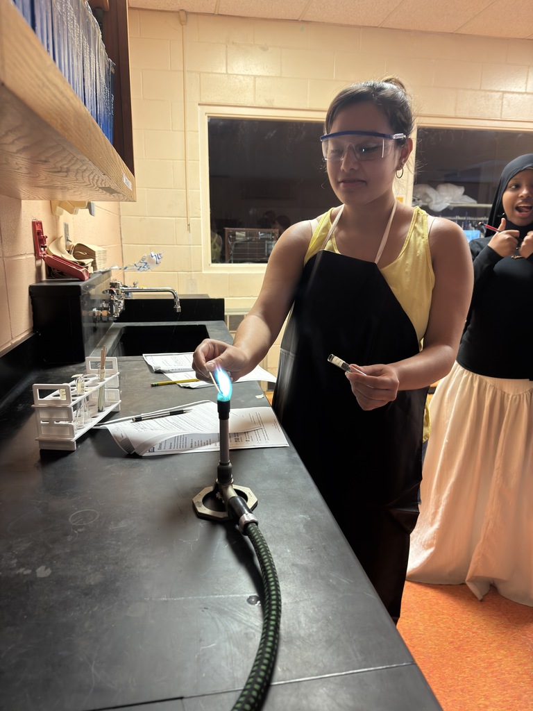 A group of students in a science classroom are gathered around lab tables wearing safety goggles. They are conducting a flame test experiment, carefully holding metal salts over a small flame. Bright colors—such as red, green, and yellow—flicker in the flames, showing the different chemical compositions. The students look focused and excited, actively participating in the hands-on activity under the guidance of Mrs. Ramos.